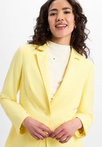 Gelber Blazer mit weicher Textur, einknopfverschluss und Reverskragen, getragen über einem hellen kremfarbenen Oberteil mit kleinen Verzierung.