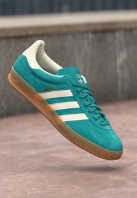 Teal Adidas Gazelle kedas ar baltajiem svītriņām un gumijas zoli, parādīti gaisā virs teksturētas virsmas.