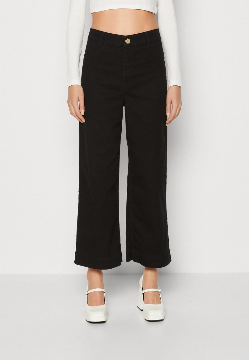 Femme portant un pantalon noir taille haute à jambes larges, associé à des chaussures Mary Jane blanches à plateforme et un top court blanc.