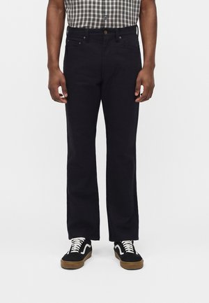 Mężczyzna w czarnych spodniach o prostych nogawkach, czarno-białych trampkach i krótki rękaw koszuli w czarno-białą kratkę, stojący na białym tle.