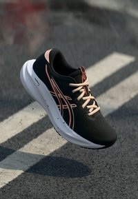 Μαύρο και ροζ αθλητικό παπούτσι τρεξίματος με λευκή σόλα, κρεμασμένο πάνω από άσφαλτο με λευκές γραμμές.