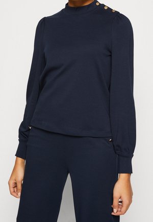Marineblaues Langarmshirt aus weichem Stoff, mit hohem Kragen, gepufften Schultern und goldenen Knopfakzenten an den Schultern und am seitlichen Saum.