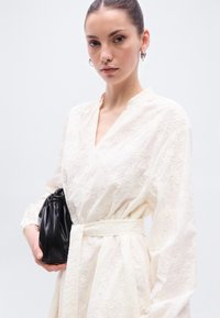 Λευκό φόρεμα από δαντέλα με μανίκια καμπάνα, εφαρμοστή μέση και κεντητό ανθισμένο μοτίβο. Μαύρο δερμάτινο τσαντάκι με λεπτομέρεια ρυτιδώσεων.