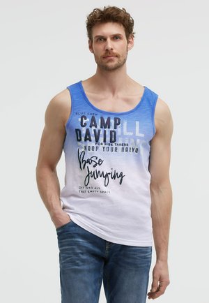 Mann mit lockigem Haar, der ein blau-weißes ärmelloses Tanktop mit Text und blaue Jeans trägt, steht mit einer Hand in der Tasche.