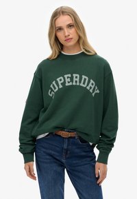 Groene oversized sweatshirt met "SUPERDAY" in gebogen grijze letters. Heeft een geribbelde kraag en manchetten, gedragen met blauwe jeans en een geweven riem.