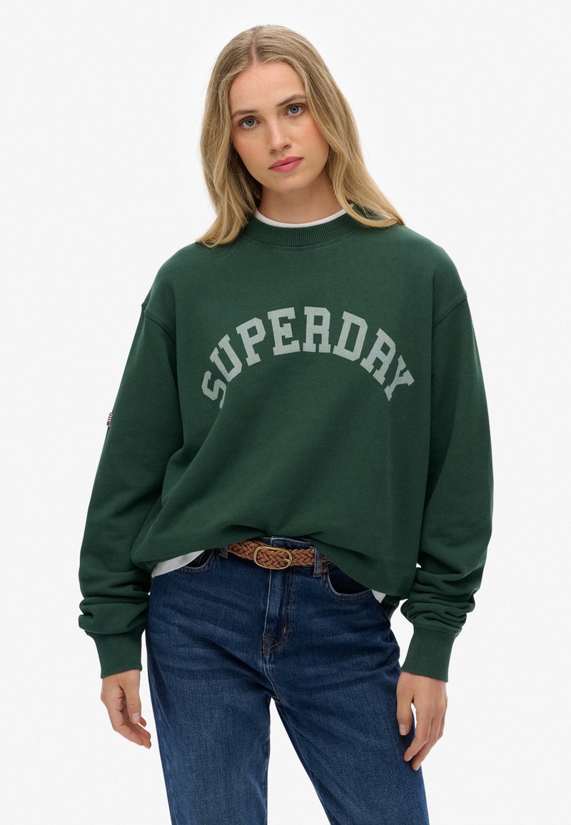 Groene oversized sweatshirt met "SUPERDAY" in gebogen grijze letters. Heeft een geribbelde kraag en manchetten, gedragen met blauwe jeans en een geweven riem.