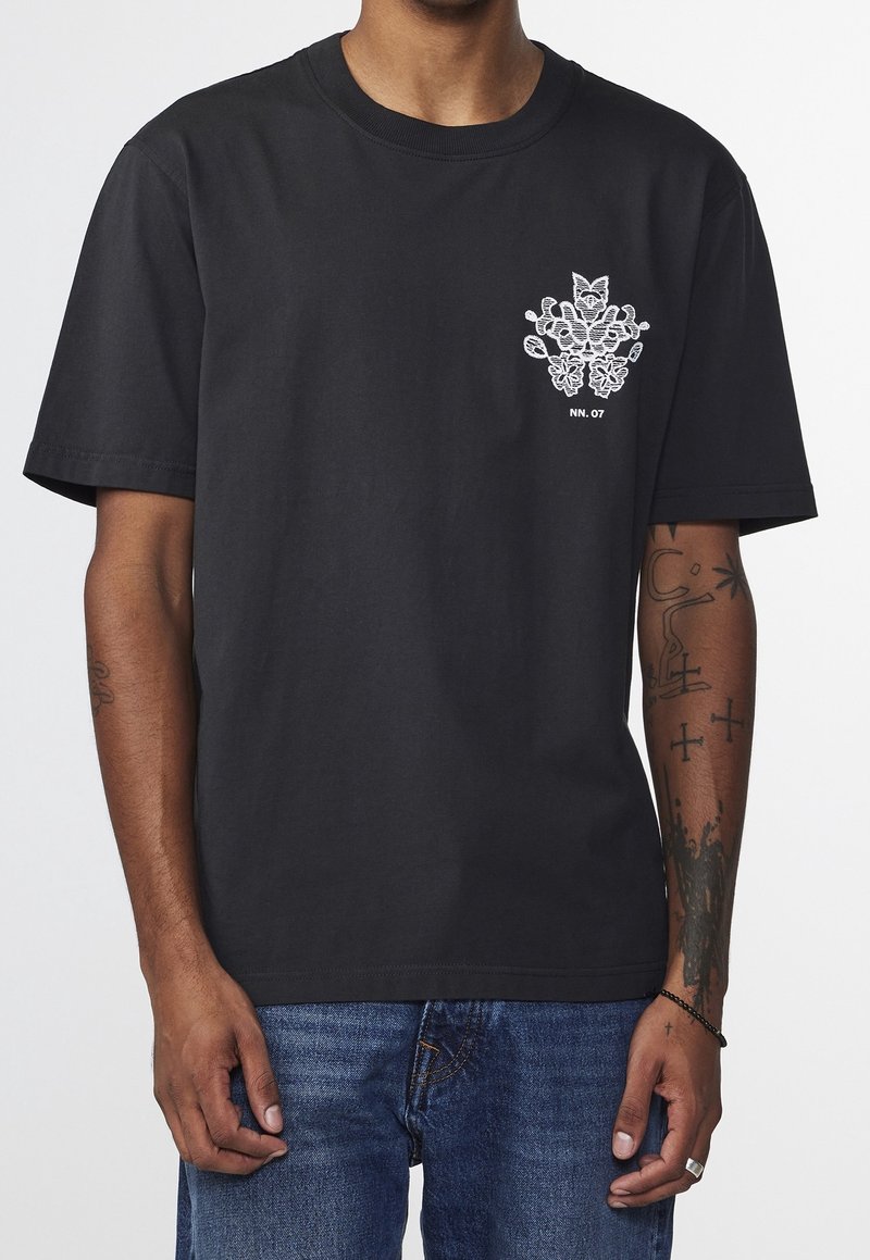 Black cotton t-shirt with short sleeves, featuring a white graphic design on the front and a crew neckline. Model wears blue jeans.