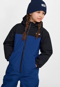 Mono de invierno azul y negro con cierre frontal, puños elásticos y bolsillos. Un gorro de punto azul marino acentúa el conjunto.