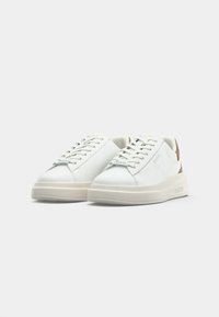 Paire de baskets en cuir blanc avec semelles épaisses beige et lacets, estampillées "Guess" sur le côté et la languette, présentées sur fond gris clair.