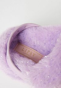 Violeta pūkaina čīpa ar mīkstu tekstūru, spīdīgiem akcentiem un lavandas krāsas siksniņu. Uz tās ir rozā etiķete ar uzrakstu "LIPSY LONDON."