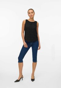 Czarny bezrękawnik z teksturowanym okrągłym wzorem, w połączeniu z niebieskimi dżinsami do kolan i czarnymi butami na obcasie typu slingback.