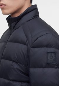Мъж, носещ черно подплатено яке с висока яка и нашивка с логото на Belstaff на горната ръкав.