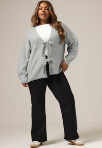 Light gray knitted cardigan with a V-neck, tie closures at the waist, long sleeves, and ribbed texture, paired with black pants and shoes.