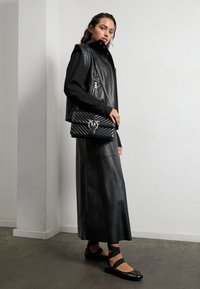 Zwarte leren vest met hoge kraag, ritssluitingen, gecombineerd met een lange zwarte leren rok en zwarte strappy slippers, met decoratieve accenten.