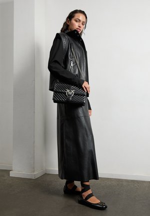 Gilet en cuir noir avec col haut, détails zippés, associé à une longue jupe en cuir noir et des chaussures noires à brides, présentant des accents décoratifs.