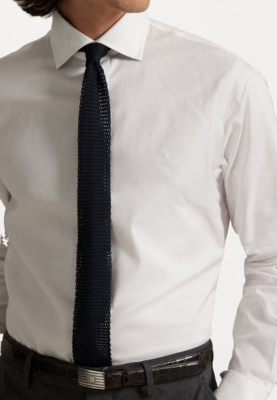 Valge särk diskreetse logoga, millele on kombineeritud tume sinine kudumismees. Riietus sisaldab tumepruunist nahkvööd hõbedase pandlaga hallides pükstes.