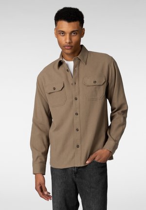 Brun, knappet skjorte i et mykt, teksturert stoff med to brystlommer og mørke knapper; båret over en hvit t-skjorte.