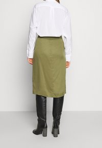 Falda verde oliva de corte recto, hecha de algodón, combinada con una camisa blanca de manga larga y botas negras de tacón alto hasta la rodilla.