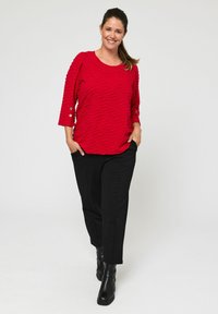Rode textuur top met een golvend patroon, driekwart mouwen en knoopaccenten, gecombineerd met zwarte broeken en enkellaarsjes.