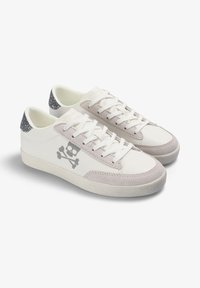 Zapatillas blancas con acentos de ante gris, suela blanca texturizada y un gráfico de calavera en el lateral. Detalles brillantes en la pestaña del talón en gris.