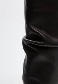 Botín de cuero negro con una textura suave y ligeramente arrugada. El diseño presenta suaves pliegues y una forma ajustada, enfatizando la flexibilidad del material.