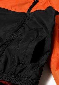 Zwarte en oranje windbreaker-jack met een ritssluiting, elastische manchetten en een soepele, lichte textuur. Inclusief zijzak.