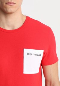 Czerwona bawełniana koszulka z krótkim rękawem z białą kieszenią, na której w czarnym kolorze znajduje się napis "CALVIN KLEIN JEANS". Okrągły dekolt.