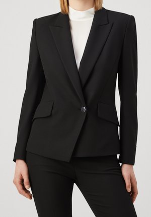 Personne portant un blazer noir ajusté avec un seul bouton et un pantalon noir assorti, sur un haut blanc à col montant.