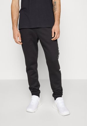 Černé tepláky s úzkým střihem nohavic, s bílým logem na boku a elastickými manžetami, doplněné o bílé sportovní boty.