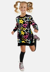 Farbenfrohes Strickkleid aus schwarzem Stoff mit leuchtenden Mustern von Sternen, Herzen und Smileys; kombiniert mit kniehohen Socken und glänzenden Sneakers.