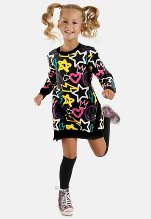 Farbenfrohes Strickkleid aus schwarzem Stoff mit leuchtenden Mustern von Sternen, Herzen und Smileys; kombiniert mit kniehohen Socken und glänzenden Sneakers.