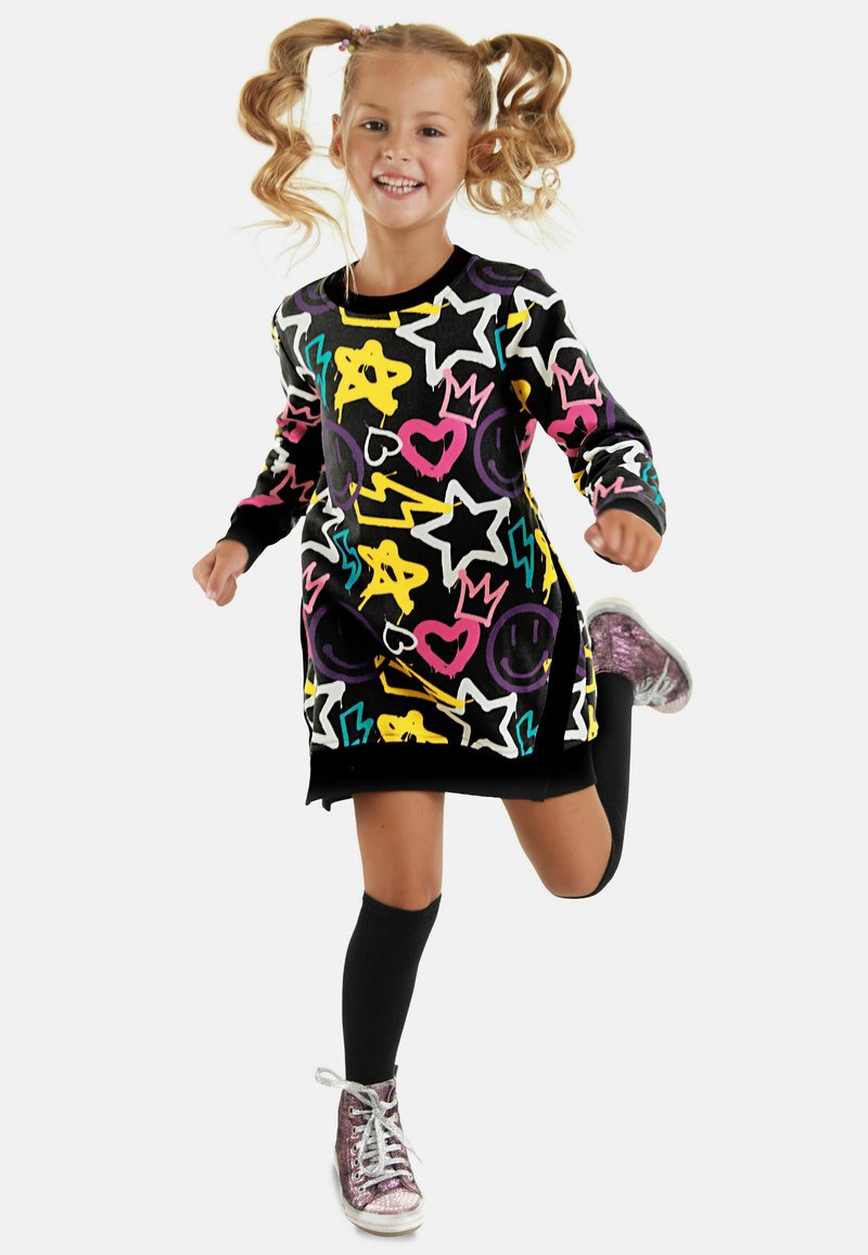 Farbenfrohes Strickkleid aus schwarzem Stoff mit leuchtenden Mustern von Sternen, Herzen und Smileys; kombiniert mit kniehohen Socken und glänzenden Sneakers.