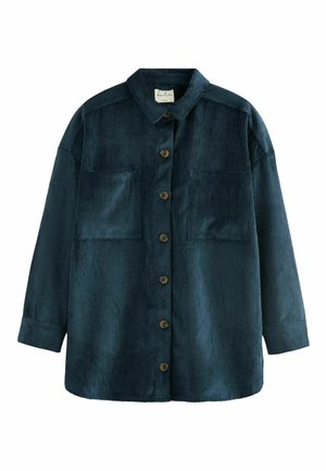 Femme portant une chemise à boutons en velours côtelé bleu foncé avec les manches retroussées et un pantalon beige clair, debout à l'intérieur avec une main dans la poche.