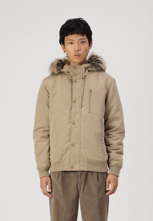 Beige puffer jacket with a faux fur-lined hood, zip and snap-button closure, and ribbed cuffs. Features one zippered chest pocket.