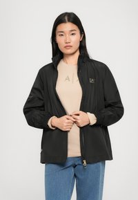 Zwarte windbreaker jas met elastische manchetten en een ritssluiting aan de voorkant. Draagt over een beige top met een gouden logo. Glad, lichtgewicht materiaal.