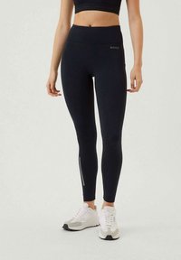 Nainen, joka on pukeutunut mustiin korkeavyötäröisiin legginsseihin, joissa on pieni logo ja heijastinraita, sekä valkoisiin ja harmaisiin lenkkitossuihin, seisoo tasaisella taustalla.