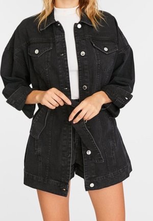 Kvinde iklædt sort denimjakke og matchende shorts, spænder bælte over hvid top, foran ensfarvet baggrund.