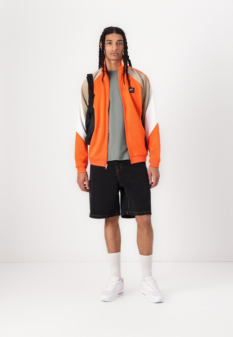 Orange Zip-up-Jacke mit beige- und weißen Akzenten, kombiniert mit einem grauen T-Shirt und schwarzen Denim-Shorts. Weiße Sneakers und graue Socken runden den Look ab.