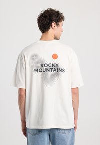 Vaaleankerman värinen t-paita, jossa on rento istuvuus, musta viivasuunnittelu ja "ROCKY MOUNTAINS" -teksti, jonka takana on oranssi ympyrä.