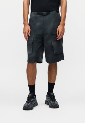 Άνδρας με μαύρα cargo σορτς με μεγάλες πλαϊνές τσέπες, μαύρα αθλητικά παπούτσια και μαύρες κάλτσες μέχρι τον αστράγαλο, στέκεται μπροστά σε απλό λευκό φόντο.