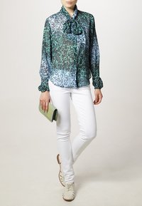 Blouse transparente à imprimé léopard en vert et bleu, avec un col noué. Associée à un pantalon blanc ajusté et des chaussures claires. Tenant une pochette verte.