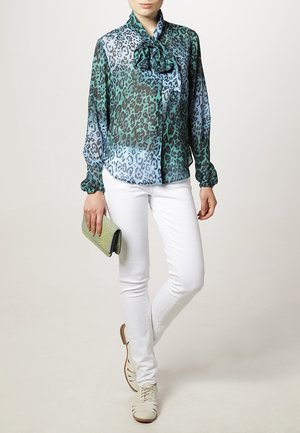 Sheer, leopard-print blouse in green and blue, with a bow tie collar. Paired with white fitted pants and light-colored shoes. Holding a green clutch.