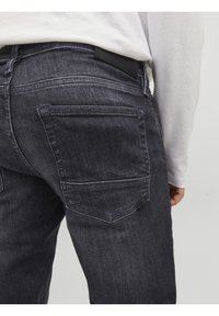 Jeans de denim negro con una textura desgastada, que presentan dos bolsillos traseros y un parche de cuero en la cintura. Diseño sencillo de corte recto.