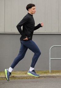 T-shirt de course à manches longues noir, pantalon ajusté bleu marine et chaussures de sport gris-vert. Accents réfléchissants sur les pantalons ; matériaux texturés.