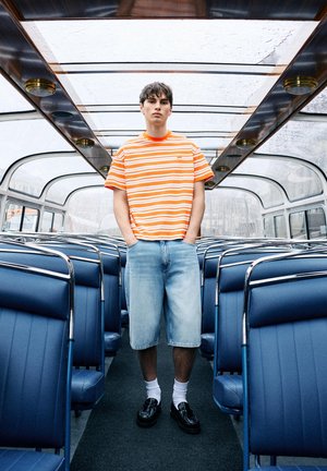 Jonge man in oranje gestreept shirt en spijkerbroekshorts staat in een lege bus met blauwe zitplaatsen, een glazen dak en metalen frames.