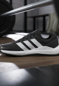 Chaussure de running Adidas noir et blanc sur le sol de la salle de sport avec une barre d'haltères en arrière-plan flou.