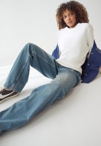 Felpa bianca abbinata a jeans in denim azzurro chiaro, dal taglio rilassato, e scarpe da ginnastica marrone scuro con accentature bianche, seduto su uno sfondo neutro.