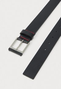 Črni usnjeni pas s srebrno pravokotno zaponko, rdeškim šivom blizu zaponke in več luknjami za nastavitev na svetli podlagi.