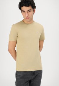 T-shirt in cotone beige con collo rotondo, maniche corte e un piccolo logo sul petto. Abbinato a jeans di denim scuri.