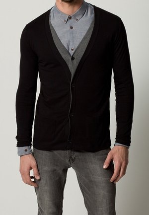 Cárdigan negro con cuello en V y botones frontales, llevado sobre una camisa azul claro abotonada. Cuenta con mangas largas y dos bolsillos delanteros.